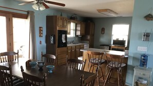 A dining area with a wooden table and chairs, a kitchen with wooden cabinets, a ceiling fan, and a small side table with decorative items. - Mother Ocean, Paradise, Oceanfront Panoramic Ocean Views, Private Beach (Holly Ridge)