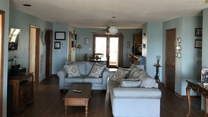 A living room with a sofa, ottoman, and coffee table, wooden flooring, and a ceiling fan. - Mother Ocean, Paradise, Oceanfront Panoramic Ocean Views, Private Beach (Holly Ridge)