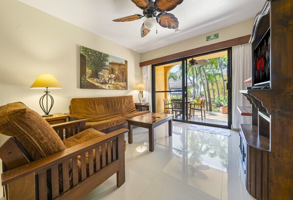 A spacious living area with a sofa, coffee table, and a television. There is a ceiling fan, a painting on the wall, and a view through the glass door.