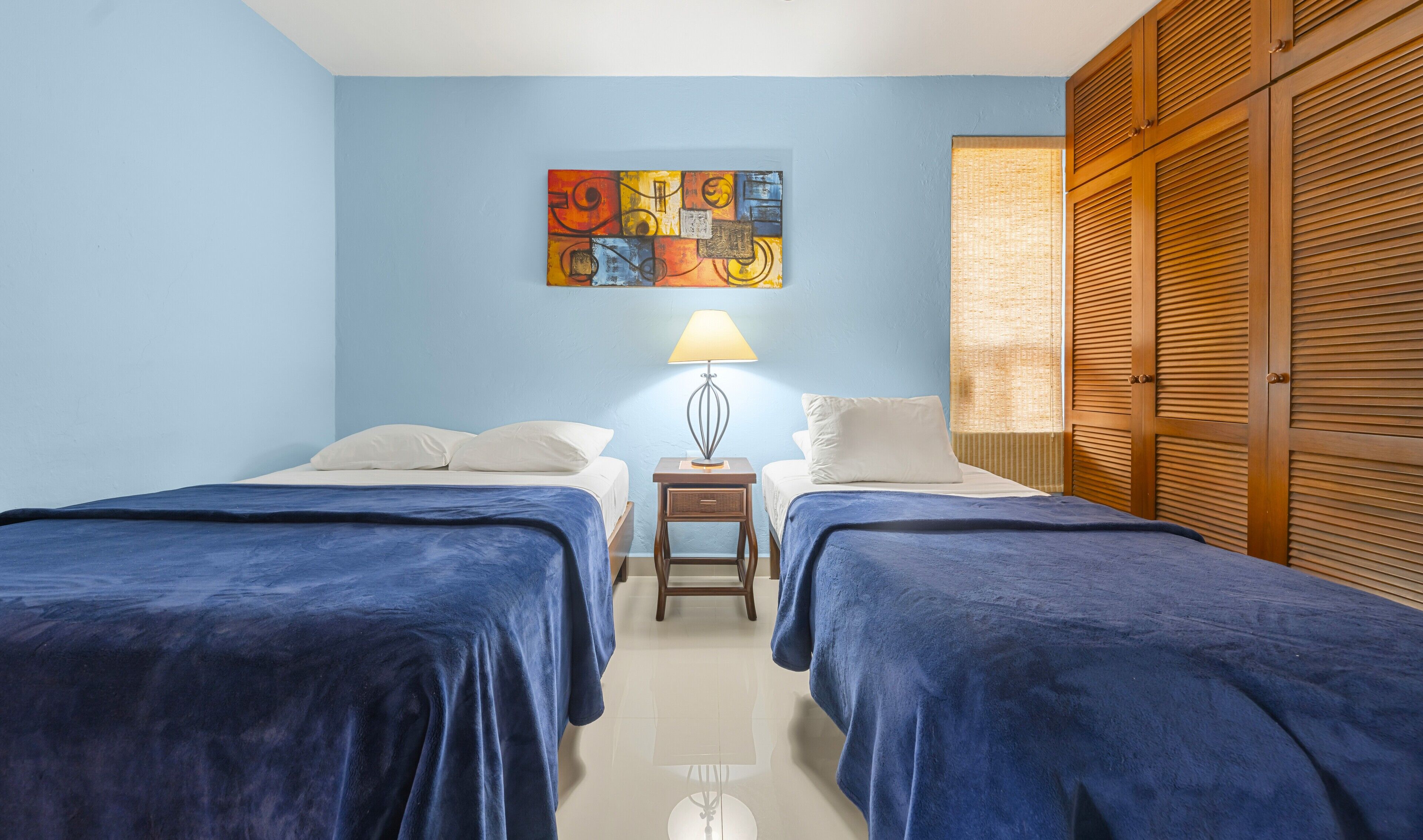 A room with two beds, a painting on the wall, a lamp, and wooden shutters.