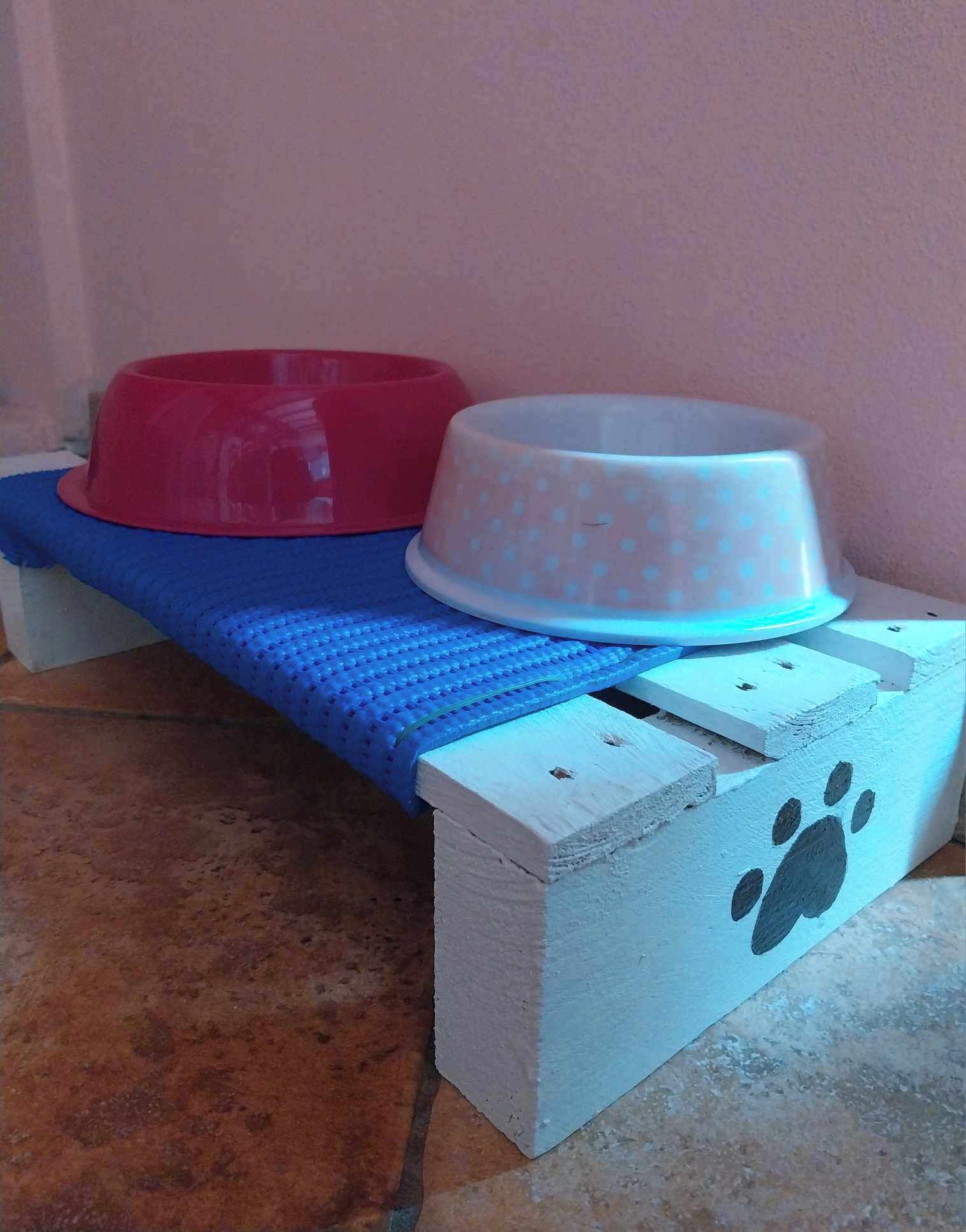 A pet bed with a blue mat and two bowls, one red and one white, on a tiled floor.