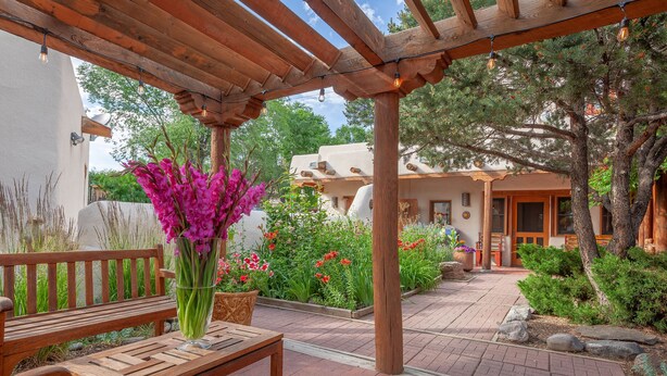 Un banco de madera con un jarrón de flores, una pérgola con luces colgantes y un jardín con diversas plantas y un edificio al fondo.