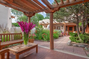 A wooden bench with a vase of flowers, a pergola with string lights, and a garden with various plants and a building in the background. - Come stay with us at La Posada de Taos ! Enjoy a unique and cozy space.  (Taos)
