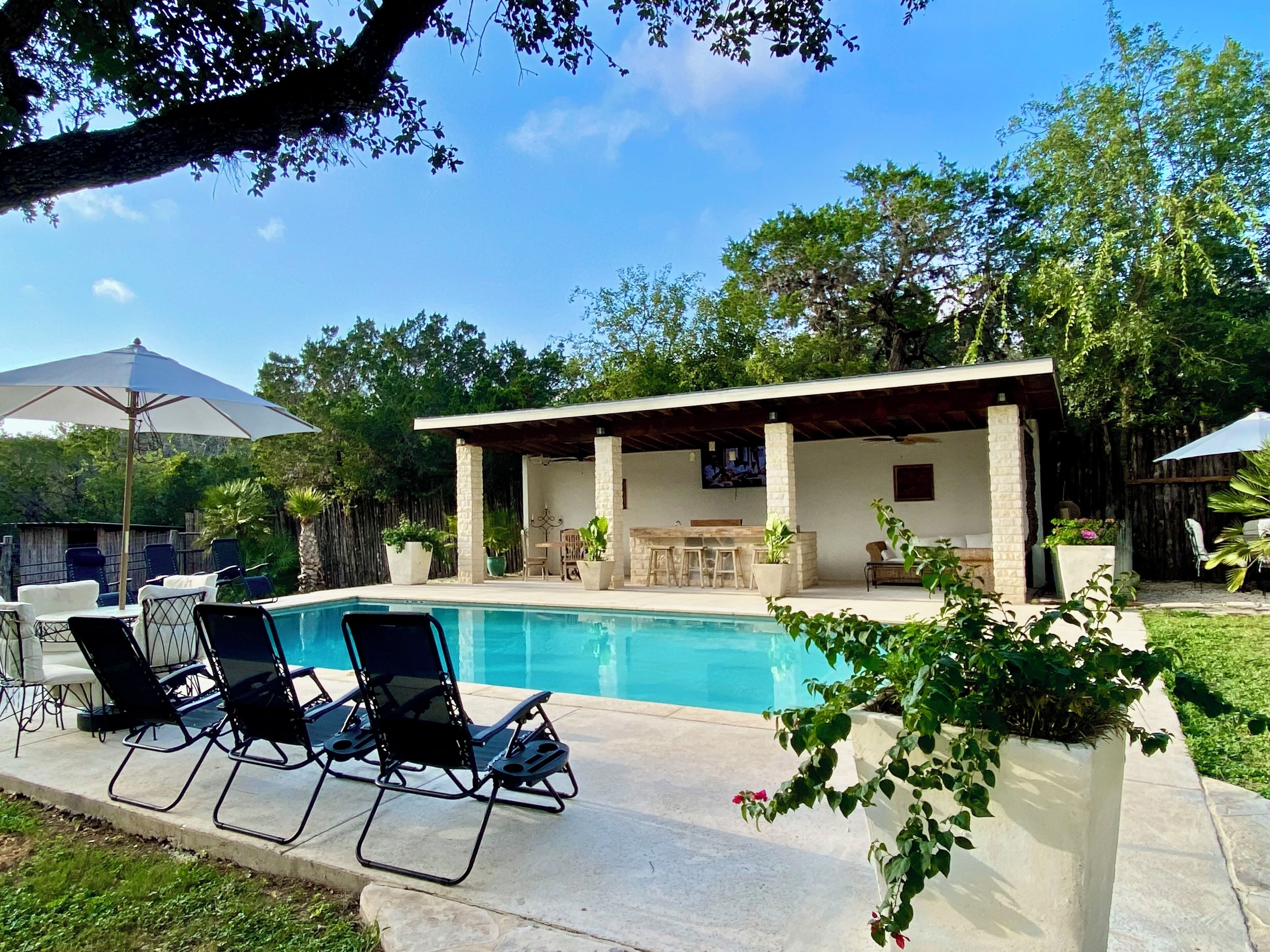 A backyard with a swimming pool, lounge chairs, a covered patio, and a TV screen.