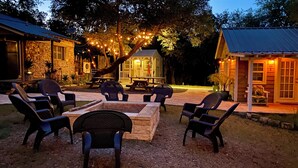 An outdoor patio area with stone seating and a fire pit, surrounded by trees and illuminated by string lights. - Historic Gated Estate, Pool, Cabana, 2 Kitchens, Lush, Kayaks, Walk 2 Frio! (Concan)