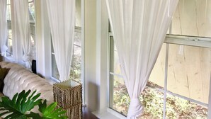A room with large windows covered by white curtains, a cushioned bench, and a woven basket. - Historic Gated Estate, Pool, Cabana, 2 Kitchens, Lush, Kayaks, Walk 2 Frio! (Concan)