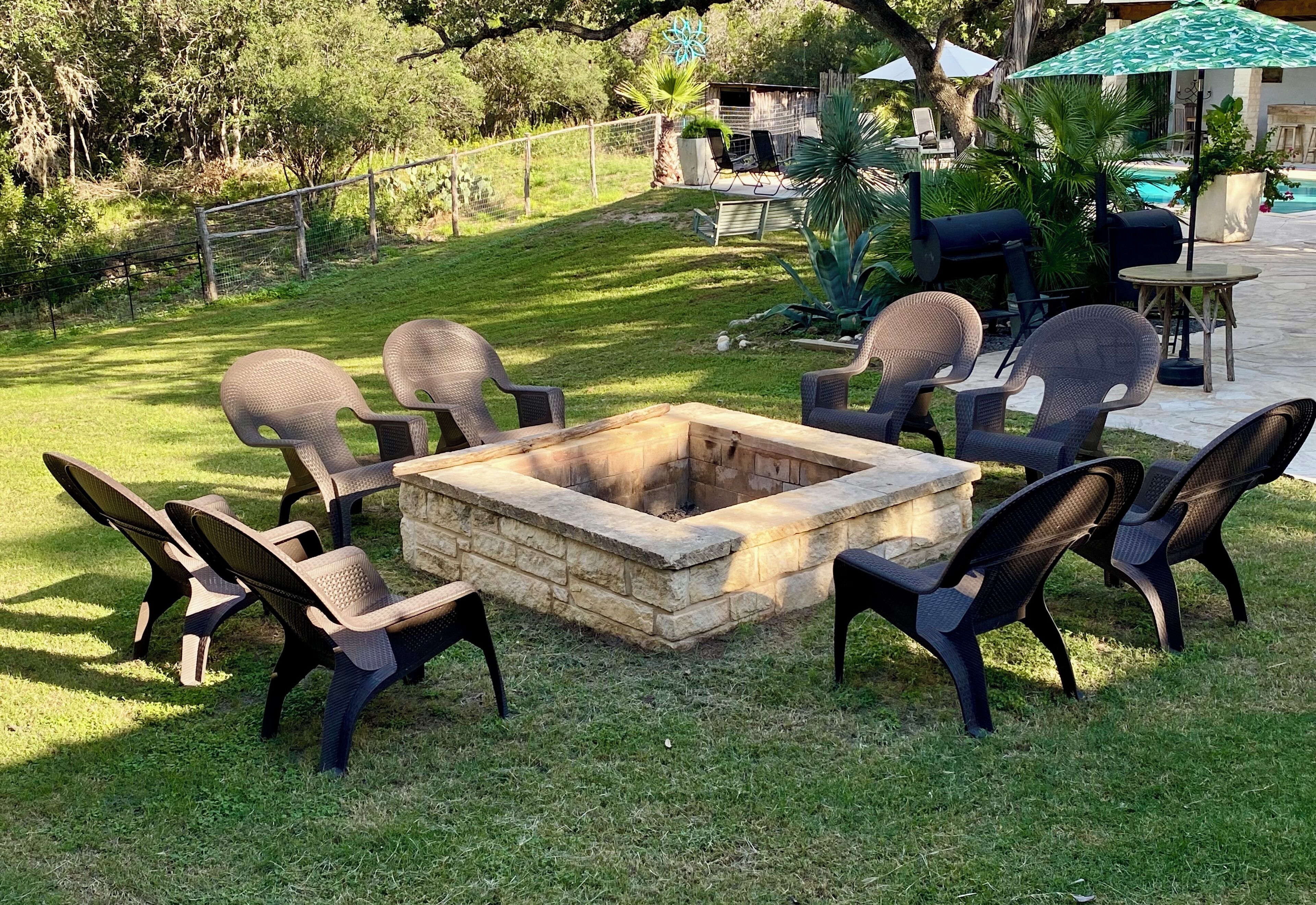 Ein Lagerfeuer, umgeben von Korbstühlen, in einem Garten mit Pool und Gartenmöbeln.