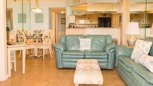 Una sala de estar con un sofá color turquesa, un reposapiés a juego y una zona de comedor con sillas blancas y una mesa.