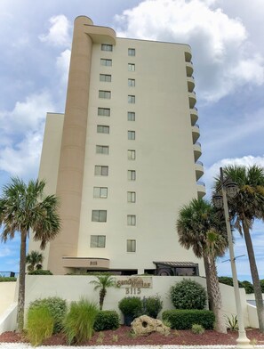 A tall, beige multi-story building with balconies, a sign reading "Sand Dollar 3115", and surrounded by palm trees and landscaped greenery. - Newly Renovated Oceanfront Pool & Deck- GORGEOUS Ocean Views!!! (Daytona Beach Shores)