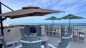 Terraza con mobiliario exterior, incluyendo sillones Adirondack y mesas, con vistas a una playa.