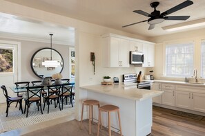 Une cuisine moderne avec des meubles blancs, un îlot central doté de tabourets, un ventilateur de plafond et un coin repas avec une table et des chaises en verre.