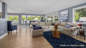 A spacious living room with a large sectional sofa, a glass coffee table, and a view of a lake. - Oasis on Murray Lake, just NE of Grand Rapids (Lowell)