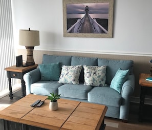 Uma sala de estar com um sofá azul claro, duas almofadas estampadas, uma mesa de centro de madeira com uma planta, um abajur e um quadro com uma imagem de um farol na parede.