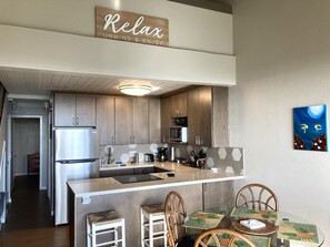 A modern kitchen with a central island, wooden cabinets, and a dining area with a glass table and wicker chairs. - Ocean View, Fully Remodeled, New AC, Walk to Beach (Kihei)