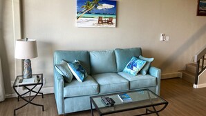 Un salon avec un canapé bleu clair, une table basse en verre et un tableau représentant une scène de plage accroché au mur.