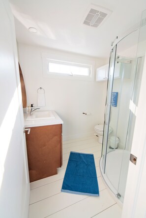A bathroom with a glass shower enclosure, a white sink, a brown cabinet, and a blue bath mat. - Floating Cottage on The LaHave River close to beaches, golf, on Lighthouse Route (Pleasantville)