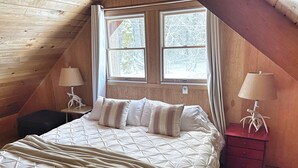 A cozy bedroom with a wooden sloped ceiling, a large bed with a quilted cover, two bedside lamps, and a nightstand with a lamp. - Cozy Riverfront Cabin Near Crystal Mountain & Sunrise Mt. Rainier National Park (Greenwater)