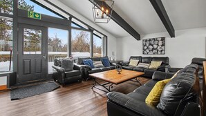 A spacious living area with a wooden floor, black leather sofas, a coffee table, and large windows offering a view of the outdoors.