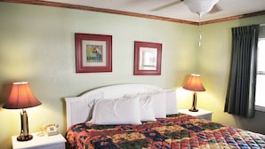 Uma cama bem arrumada com um edredom colorido, duas luminárias de cabeceira, uma mesa de cabeceira branca e duas fotos emolduradas na parede.