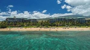 Ein Strandhotel mit mehreren Gebäuden, einem Sandstrand und klarem türkisblauem Wasser.