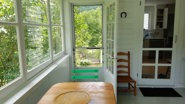 Ein Zimmer mit Holztisch, einer Bank und Blick auf Grün hinter großen Fenstern.