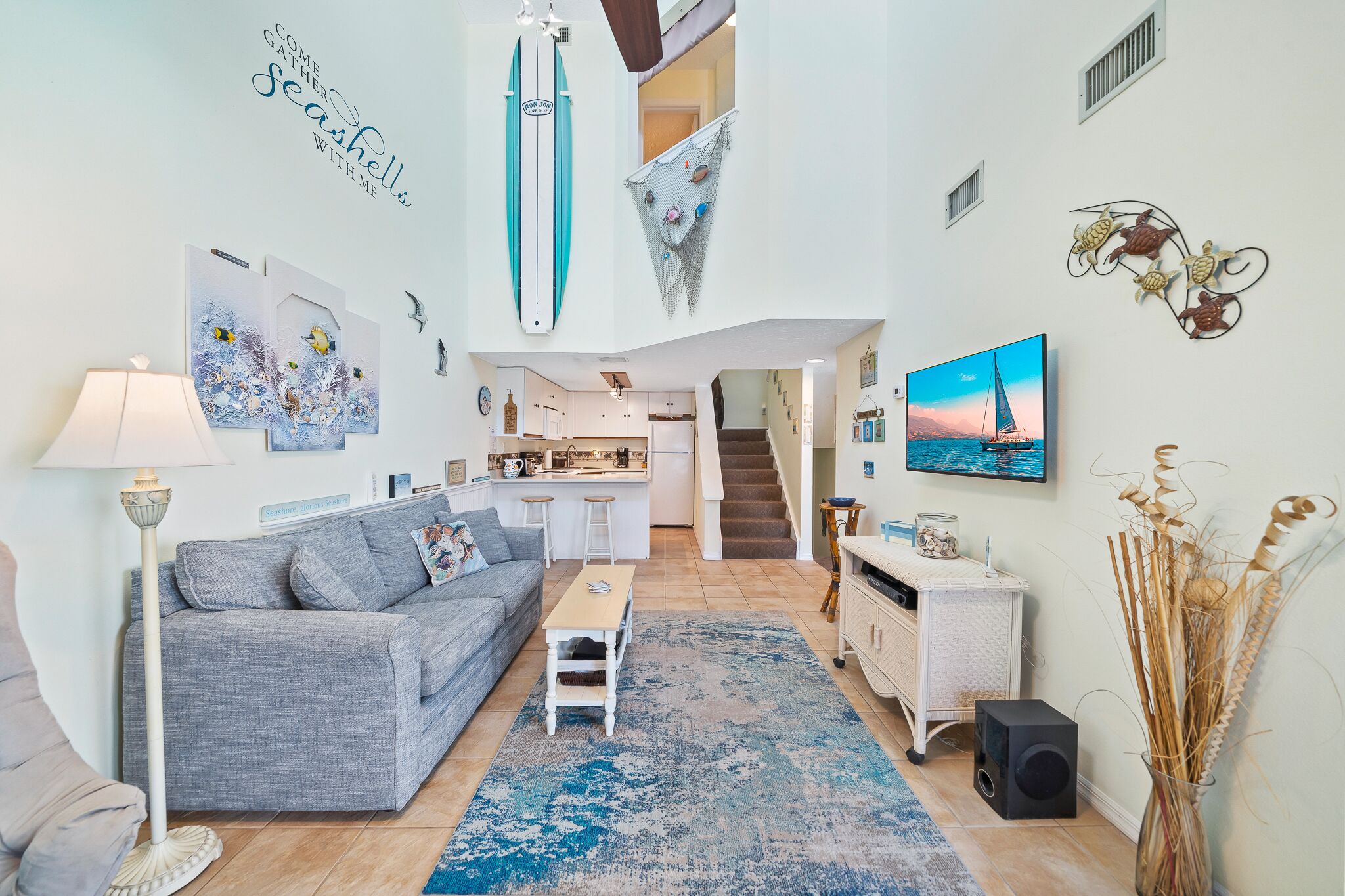 Een ruime woonkamer met een grijze bank, een bijzettafeltje en een aan de muur bevestigde televisie. Achtergrond is een keuken, een trap leidt naar een hoger gelegen verdieping, en er is decoratieve wandkunst, waaronder een surfplank en zeedieren.