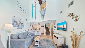 Ein großzügiger Wohnbereich mit grauem Sofa, Couchtisch und an der Wand montiertem Fernseher. Im Hintergrund befindet sich eine Küche, eine Treppe führt auf eine obere Ebene, und an den Wänden sind dekorative Elemente zu sehen, darunter ein Surfbrett und Meereslebewesen.