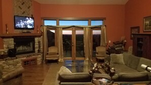 A living room with a stone fireplace, a television, a glass door leading outside, and a sofa. - Spacious High Country Custom Home with Stunning Views (Williams)