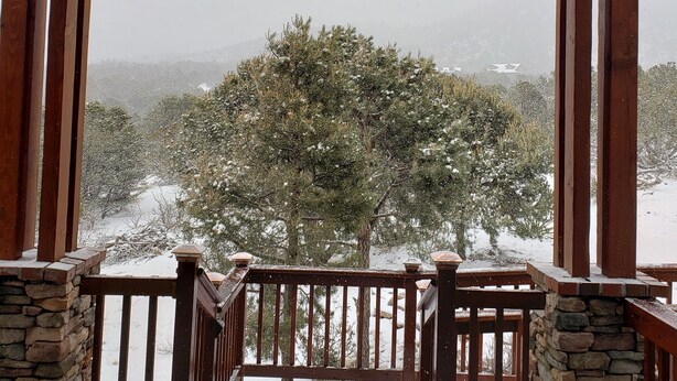 A covered outdoor area with a stone wall and wooden railing, leading to a snowy landscape with trees. - Spacious High Country Custom Home with Stunning Views (Williams)