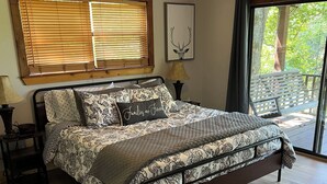 Une chambre à coucher avec un lit, un plancher en bois, un ventilateur de plafond, des stores aux fenêtres et une vue sur l’extérieur.