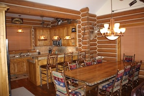 Cozy cabin interior with a central dining table and six chairs, surrounded by wooden cabinets and pendant lighting.