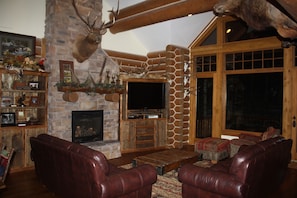 Cozy living room with a stone fireplace, leather sofas, and a mounted deer head, featuring a large window and wooden beams.