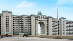 A large, multi-story hotel with a central arched entrance, numerous balconies, and a parking area with a few cars.