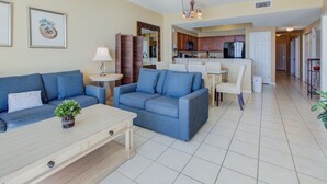 A living room with a blue sofa, a coffee table, and a small table with a lamp. There is a kitchen area with cabinets and a dining table in the background.
