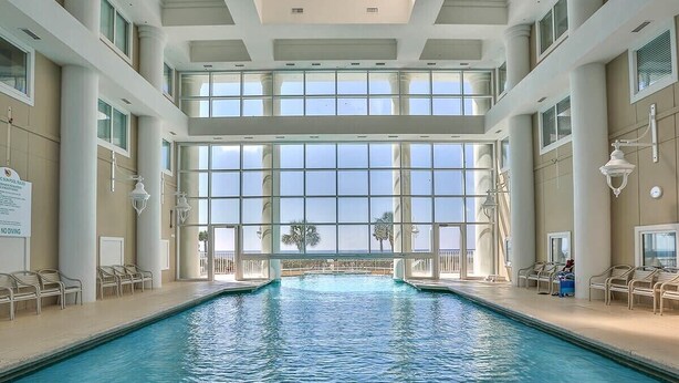 An indoor swimming pool with clear glass walls, multiple lounge chairs, and a view of palm trees and a balcony.