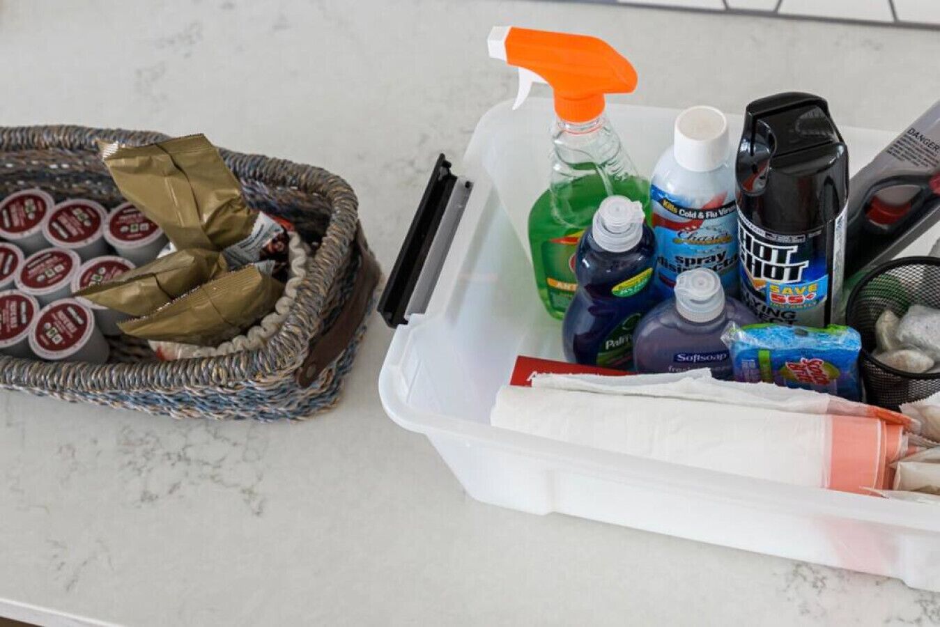 En köksbänk med en korg full av inpackade varor, en plastbehållare med rengöringsmedel och en flaska diskmedel.