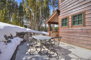 Una cabaña de madera con un patio que tiene mesas y sillas de metal, rodeada de nieve y altos pinos.