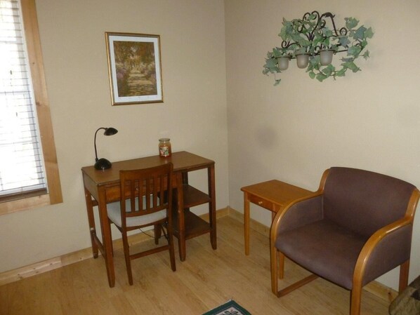 A room with a wooden desk, a chair, a small table, and a purple armchair. There is a picture on the wall and a plant on a metal stand.