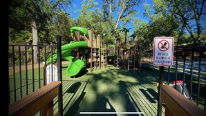 Ein Spielplatz mit einer grünen Rutsche und Klettergerüsten, umgeben von Bäumen und einem Schild mit der Aufschrift 'Hunde verboten'.