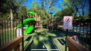 Um parquinho com escorrega verde e estruturas de escalada, cercado por árvores e uma placa indicando 'Não são permitidos cães'.