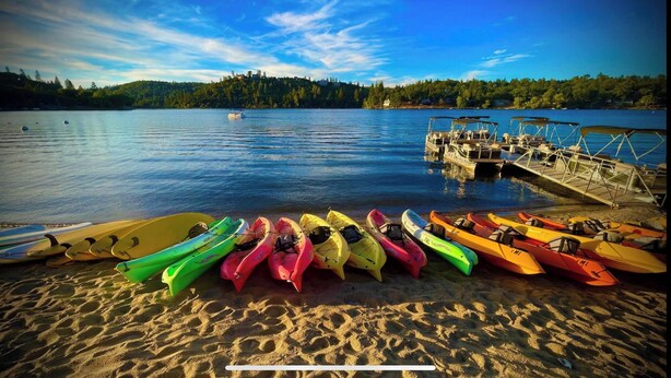 Eine Reihe bunter Kajaks an einem sandigen Strand, im Hintergrund ein Steg und Boote.