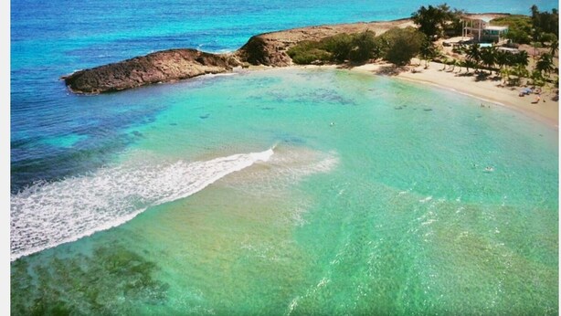 Une vue sur la côte, avec une eau turquoise cristalline, un promontoire rocheux et une plage bordée de palmiers et d’un bâtiment.