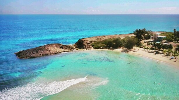 Vista costera con aguas turquesas cristalinas, un promontorio rocoso y una playa con palmeras y un edificio.