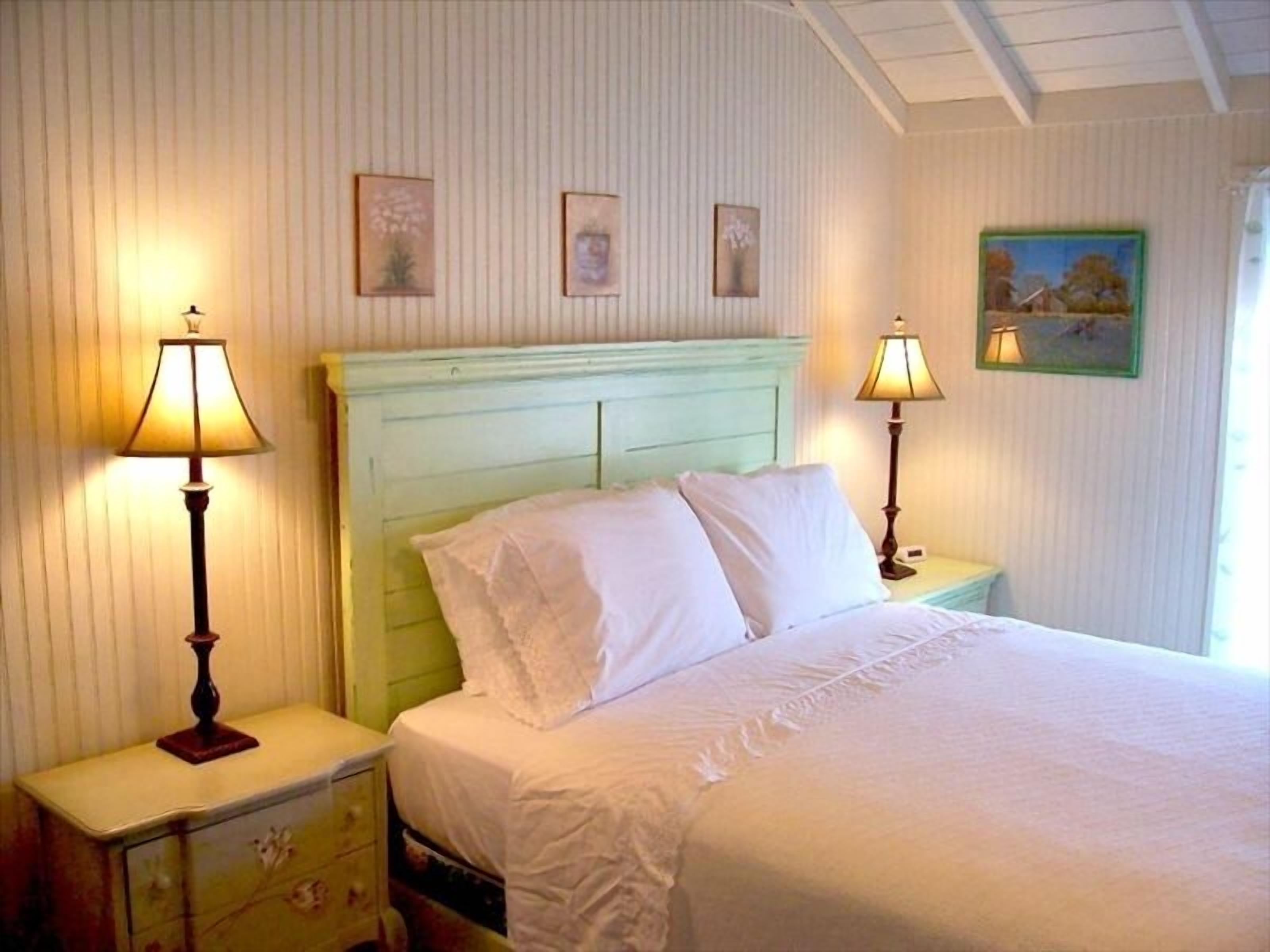 Una habitación con una cama, dos mesitas de noche con lámparas, un aparador y tres cuadros en la pared.