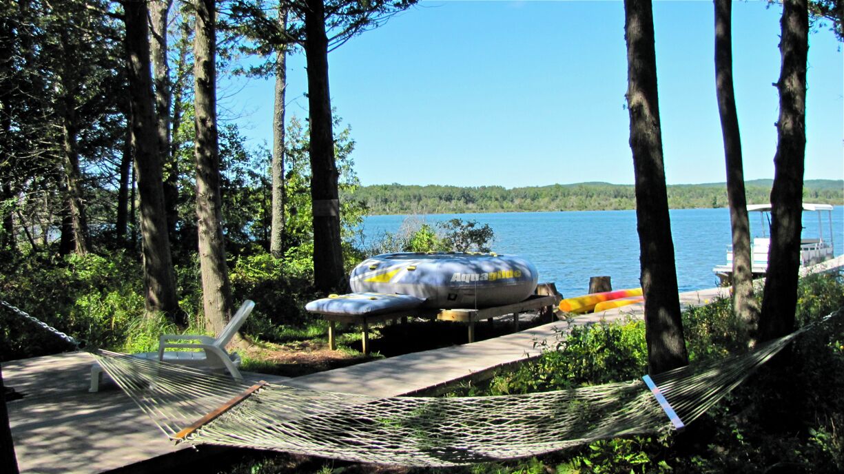 Un hamac et un espace abrité avec un bateau amarré au bord d’un lac, le tout entouré d’arbres.