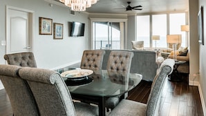 A dining area with a glass table and tufted chairs, a ceiling fan, and a view of the ocean through large windows.