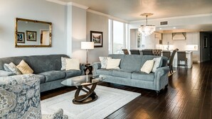 A modern living room with a grey sofa, a coffee table, and a patterned armchair. There is a dining area with a chandelier and a kitchen in the background.