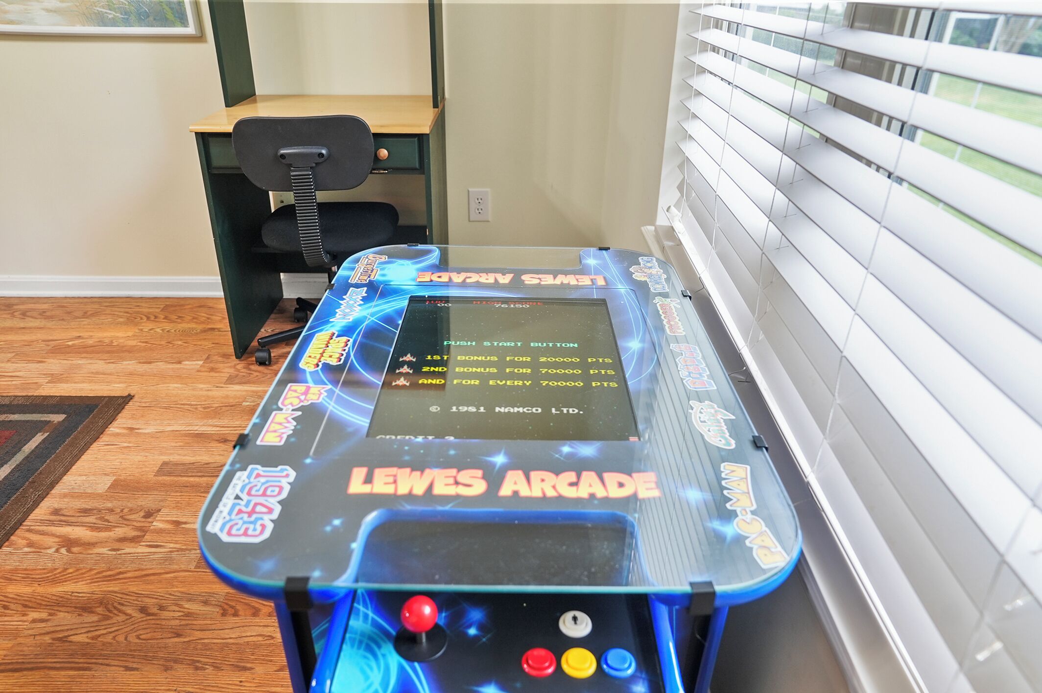 Een spelconsole met scherm en knoppen, een bureau, een stoel en een raam met jaloezieën.