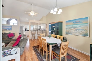 A modern living room with a sofa, dining table, and chairs, a kitchen in the background, and a beach-themed painting on the wall. - Prime July 4th Dates Still Available Summer Beach Getaway Near Lewes & Rehoboth (Lewes)