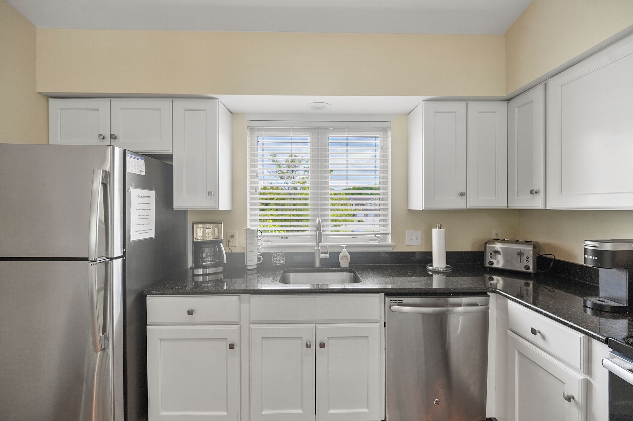 A modern kitchen with white cabinets, stainless steel appliances, a window with blinds, and a black countertop.